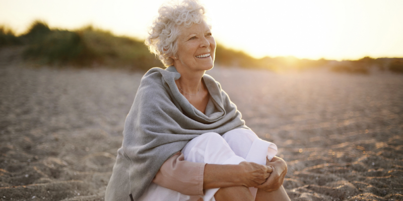 An older woman sitting on the beach, smiling with teeth whitened before her vacation.